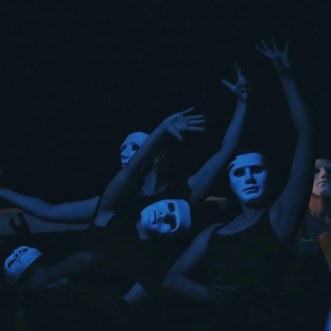 Impactante escena coral con actores enmascarados bajo luz azul en la obra "¿Qué sueña el cielo cuando somos desierto?", un ejemplo vibrante de los montajes de egreso resultantes de las clases de teatro del CEDRAM.